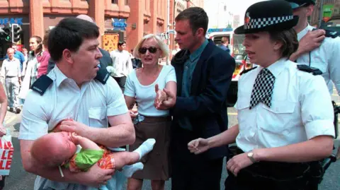 Manchester Evening News Lisa Hughes, with blonde hair and wearing sunglasses, looks visibly distressed as her seven-month-old son Sam - wearing a green t-shirt and orange checked shorts and white socks - is carried by a male police officer.