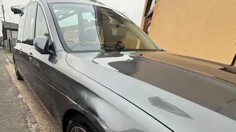 A grey Mercedes hearse parked beside a yellow wall. it has a long scratch down the side above the wheel. 
