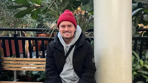 Rii Schroer Alex Warwick smiles as he sits on a bench surrounded by tropical-looking plants. He is wearing a red woolly hat, a light-grey hoody and a black jacket.