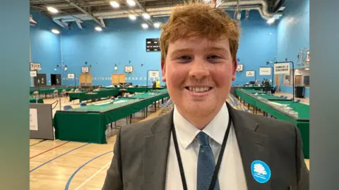 A man with red hair smiles at the camera while wearing a smart suit with a Conservative badge on his lapel