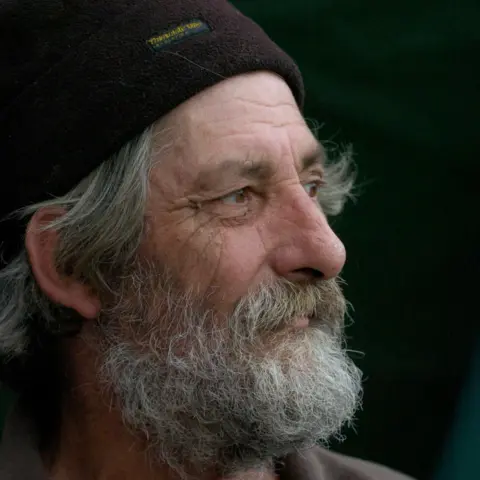 Holmes Family A man with a black woolly hat on and a grey beard looks off to the right