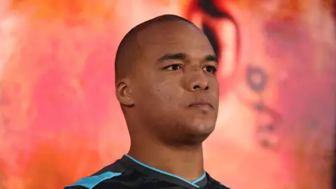 PA Media Fabio Wardley during a weigh-in at Spitalfields Market. He looks away from the camera has he stands on a stage in front of a screen. He wears a black and blue Ipswich Town football shirt. 