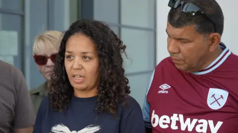 Jamie Niblock/BBC Anita Rose's eldest daughter - a woman with long curly black hair - standing outside caught. Next to her is Anita Rose's partner, who is wearing a West Ham shirt and looking over her shoulder. 