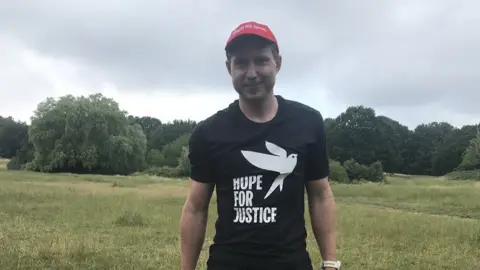 BBC Aaron Robinson, aged 40, smiles at the camera wearing a black t-shirt with the Hope for Justice's charity branding. Behind him is a grassy field where he runs.