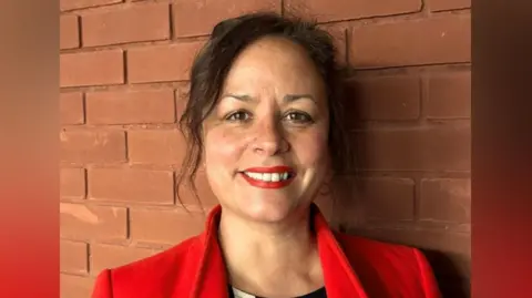 Supplied Michelle Daurat has brown hair and eyes and her hair is tied back. She is wearing a red lipstick, which matches her coat, and a black top. She is smiling.
