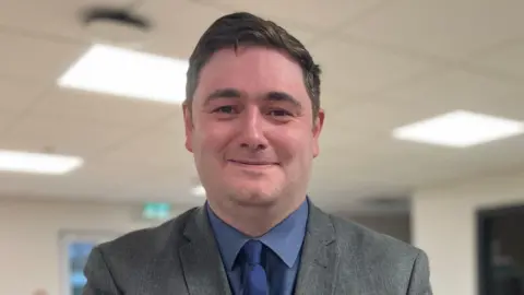 Mayor Chris Cooke has short brown hair and a grey suit and blue shirt. He has a blue tie. He is clean-shaven and standing in a classroom which is blurred out. 