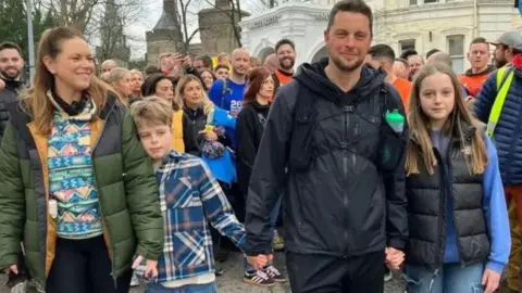 Craig holding his son and daughter's hands, with his wife to the right hand side. A crowd of people stand behind them. 