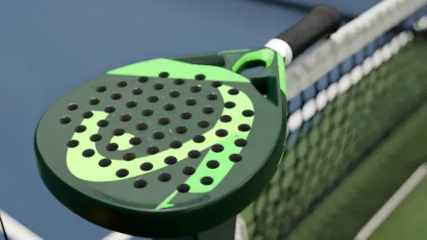 A green padel bat which has holes in it, and a black handle. It is balancing on a black net stretching across a court. The court's floor is blue with shite lines.