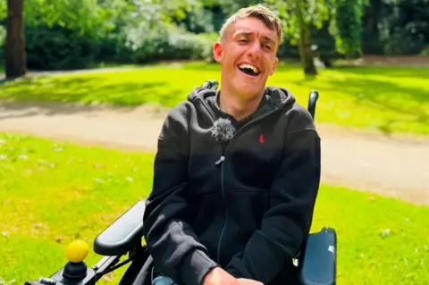 BBC Jake Smith, a young man in a wheelchair, he is laughing heartily in a green park setting.