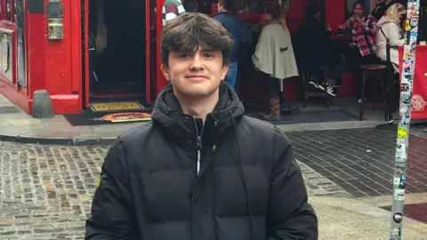 Elliot Winterbottom A man with dark hair and a black coat is looking at the camera and a street is behind him in the bottom half of the photo. People are sitting on stools outside a pub or bar in the top right of the image.