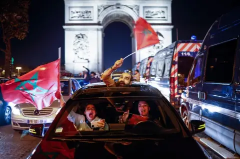 EPA People in a car waiving Moroccan flags against a backdrop of the l'arc de triomphe. They look very happy and excited.