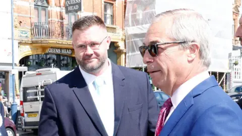 SteveFinnPhotography Taylor, wearing a suit with a light blue tie, with a street seen in the background. He is stood next to Nigel Farage, who is wearing a suit and sunglasses.