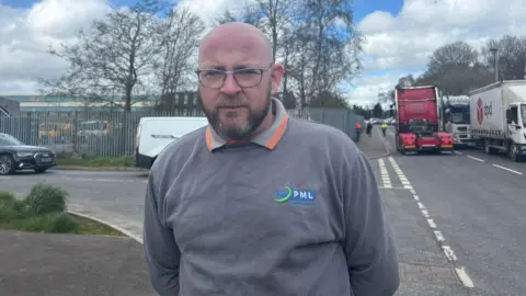A bald man with a beard and wearing glasses and a grey sweatshirt stands on a road with a number of lorries in the background. 