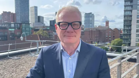 A man with white hair and dark-framed glasses, wearing a navy suit and blue shirt, stands on a city balcony. Beyond him skyscrapers and multi-storey flats can be seen.