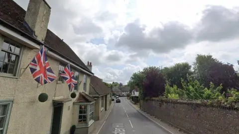 Cream building with pale green paining windows on the left. Union Jack flags hang on posts from upper floor and a road runs alongside it, top to bottom. A red brick wall is on the right side of the image