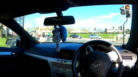 View from front seat of car looking out onto a junction. Part ofthe junction is obscured by two large stuffed toys dangling from the rear-view mirror.