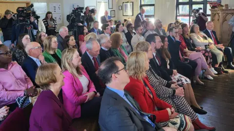 A group of Labour politicians and party members sat listening to Eluned Morgan, with Jayne Bryant, Lee Waters, Jeremy Miles and Mark Drakeford among them.