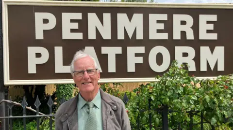 A man standing in front of a vintage style railway station sign that reads Penmere Platform. He has white hair and wears a green tie 