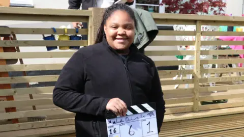 PQA Kinara wearing a black jacket, smiling and holding a black and white clapperboard. She is standing in front of some wooden decking outdoors.