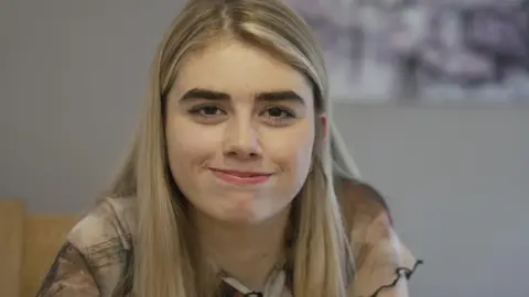 A teenage girl with blonde hair and brown eye looks directly into the camera smiling, the background is out of focus and grey.