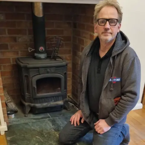 Kerry Walpole Kerry Walpole is kneeling in front of a wood burner. He's wearing a grey fleece over a dark T-shirt and jeans.