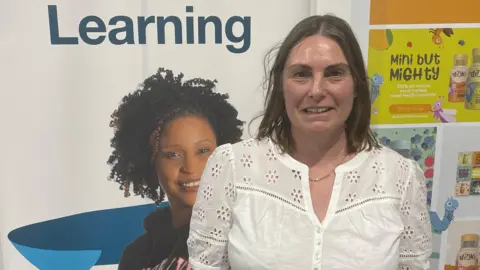 A woman smiles at the camera as she stands in front of an information board detailing adult learning. She has shoulder length brown hair and she is wearing a white blouse. 