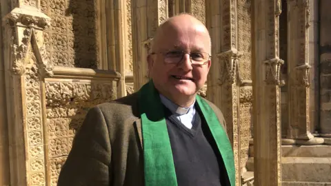 Reverend Alan Robson. He is wearing glasses, a clerical collar, a blue jumper, tweed jacket and green stole. He is standing in Lincoln Cathedral.