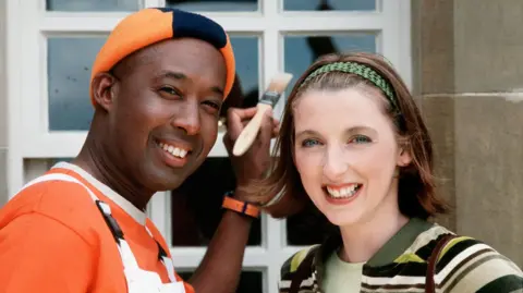 Rodd Christensen, as Spencer the Painter, and Julie Wilson Nimmo, as Miss Hoolie the nursery teacher, stand in front of a window that is being painted