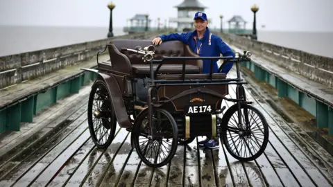 A rainy Clevedon Pier, with Mark Reber standing beside a brown wooden framed Victorian motor car