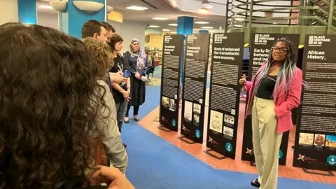 Chantelle Lunt is giving a talk at the opening of the museum exhibit. 
A crowd of people are stood listening to her. 
She is wearing a black top, pink jacket and cream trousers. 