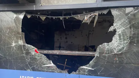 PA Picture shows a large broken hole in a window at the front of a cafe in Belfast - the picture focus on the large hole in the middle of the window, which shows most of the window gone. At the edges, are broken sharp crags of the remaining glass in the frame. Inside the cafe has been destroyed by fire. 