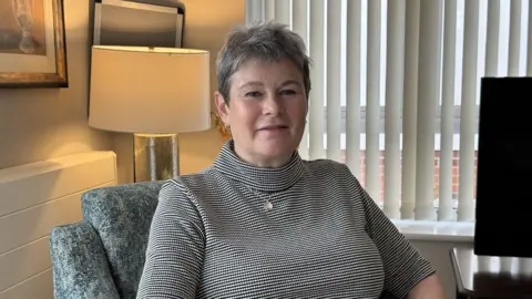 A woman with short grey hair and a grey top in a grey hair with white blinds behind her