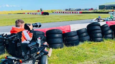 A young man in a wheelchair pointing a camera at a go-karting track