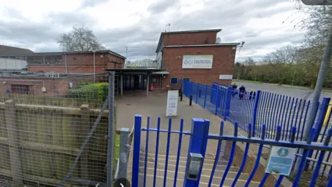 Google The outside of a school building seen from the blue metal gates with intercom. It is a two storey building with a sign stating the school name on the side. A parking area can be seem to the side, with two blurred figures standing in it.