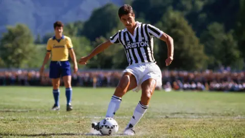 Getty Images Ian Rush, wearing a black and white striped Juventus shirt and white shorts and socks, takes a penalty as an opposition player in a yellow and blue kit watches in the background
