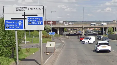 Google Road junction with overhead bridge and multiple cars, featuring directional road signs pointing to Glasgow, Kilmarnock, Cambuslang, Carlisle and nearby city centre routes.