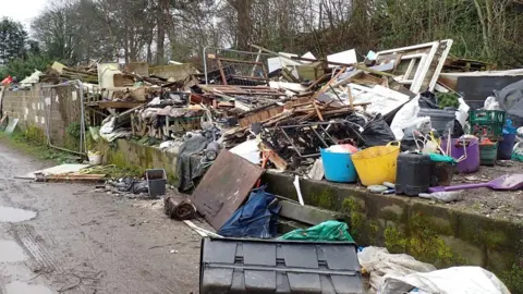 A large pile of waste including bits of wood, plastic tubs and other unidentifiable waste. Trees in the background and a lane in the foreground.