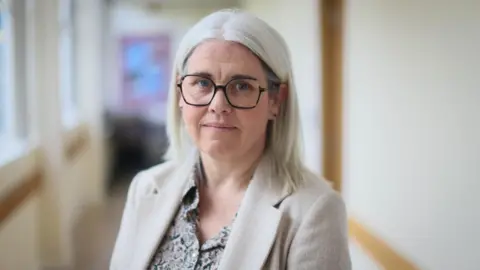 Louise Moir standing in a school corridor. She has shoulder-length, grey hair and glasses, and is wearing a cream jacket and a patterned top.