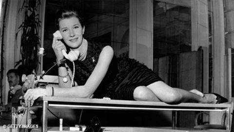 Lois Maxwell in a black dress, knees exposed, posing on top of a coffee table holding a receiver from a telephone.  A typewriter is behind her.