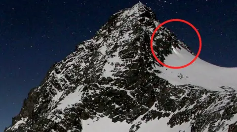 Mountain against a night sky with a red circle highlighting torchlight on the side of the mountain.