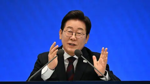 Lee Jae Myung in a suit, sitting in front of two microphones. He is speaking and gesturing with his hands.