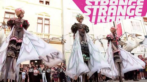 People in bird costumes on stilts and the Strange News logo