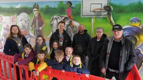 Wychavon District Council A group of people, adults and several children, are standing in an underpass in front of a wall. The wall is painted with a mural which depicts a pink pig, a white dog, Harry Potter riding a scooter, a girl riding an elephant and a basketball hoop.