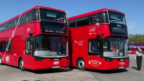 BBC A woman stands next to two red double decker buses in a bus depot.