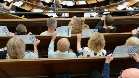 The photo is taken from above a public gallery. Several people are visible in tiered wooden rows of seating looking down from a balcony. Below is a circle of seats with more people sitting down. The people above are holding up pieces of paper.