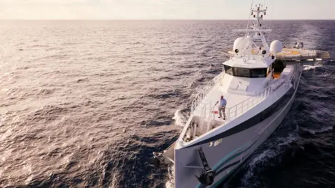 James Honeyborne A dramatic wide shot of Sir David Attenborough standing on the bow of a research vessel in the middle of the ocean on a sunny day