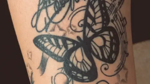 The image shows a close-up photograph of a tattoo. The design centres on a large black butterfly surrounded by musical notes.