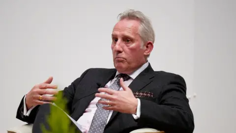 Reuters A man in a dark suit and light pink shirt and tie is sitting against a plain white wall, looking to the left of the camera. He has grey hair and a hankerchief in his breast pocket. His hands are open and in the air mid-movement while he speaks. 