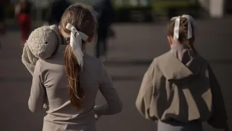 BBC Two young girls walk alongside each other with their backs to the camera. They're wearing grey with their hair tied back in a bow. The one on the left is carrying a large stuffed toy rabbit. 