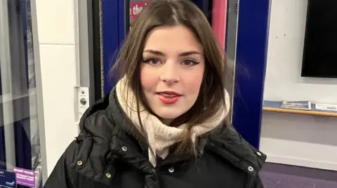 BBC/Richard Edwards A young woman with long dark hair and dark eyes. She is wearing a cream-coloured scarf and a black coat. Behind her is the blue door frame and window of a railway station waiting room